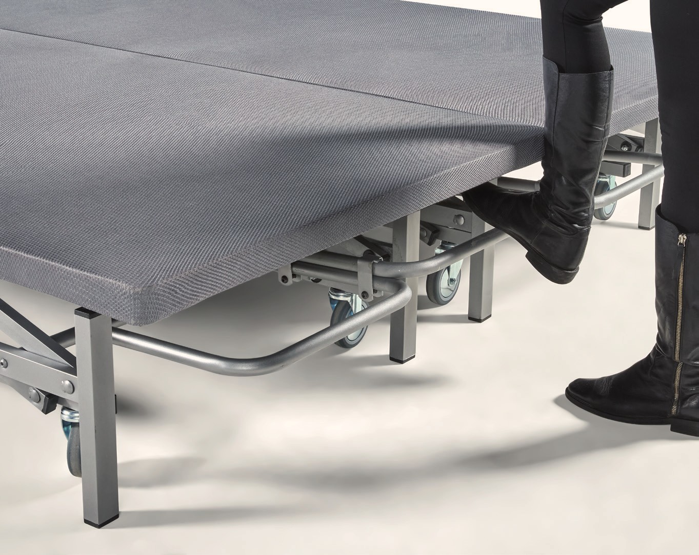 Una persona acciona con el pie el pedal de acero de una cama elevable: el tubo recorre la base y la cama sube al pisarlo.