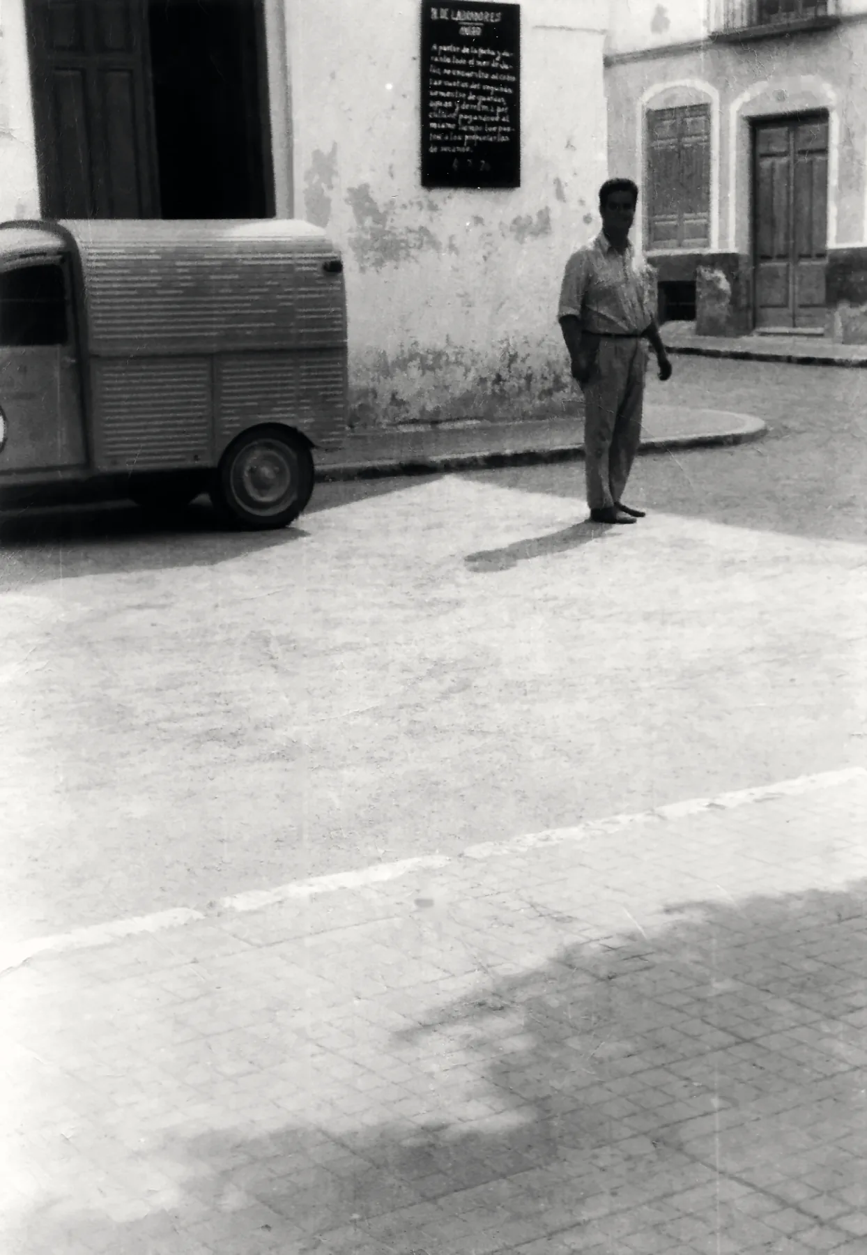 Fotografía antigua en blanco y negro: un hombre de pie en una calle de pueblo, junto a una furgoneta de chapa ondulada.