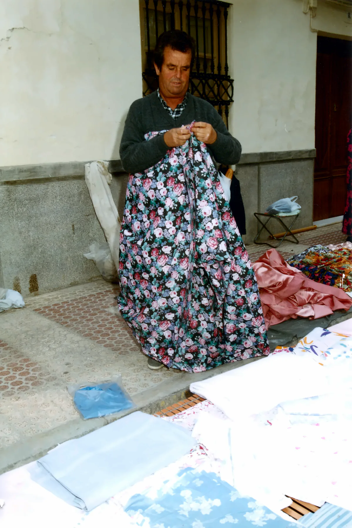 Fotografía familiar en color: un vendedor sostiene una tela estampada de flores en un puesto de mercadillo, con sábanas y tejidos extendidos en el suelo.