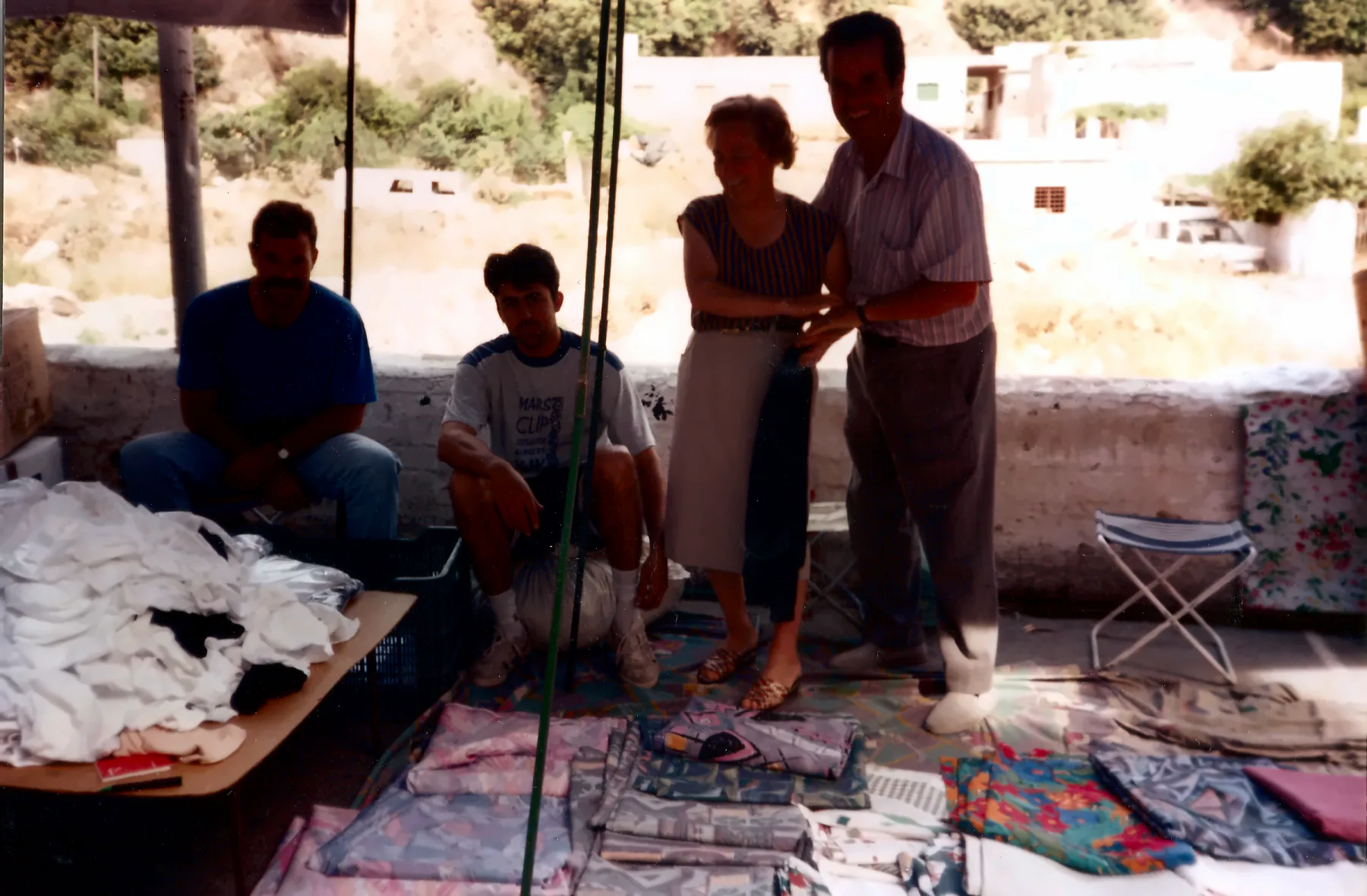 Fotografía familiar en color: cuatro personas junto a un puesto de mercadillo con ropa de cama y tejidos, en un pueblo de ladera.